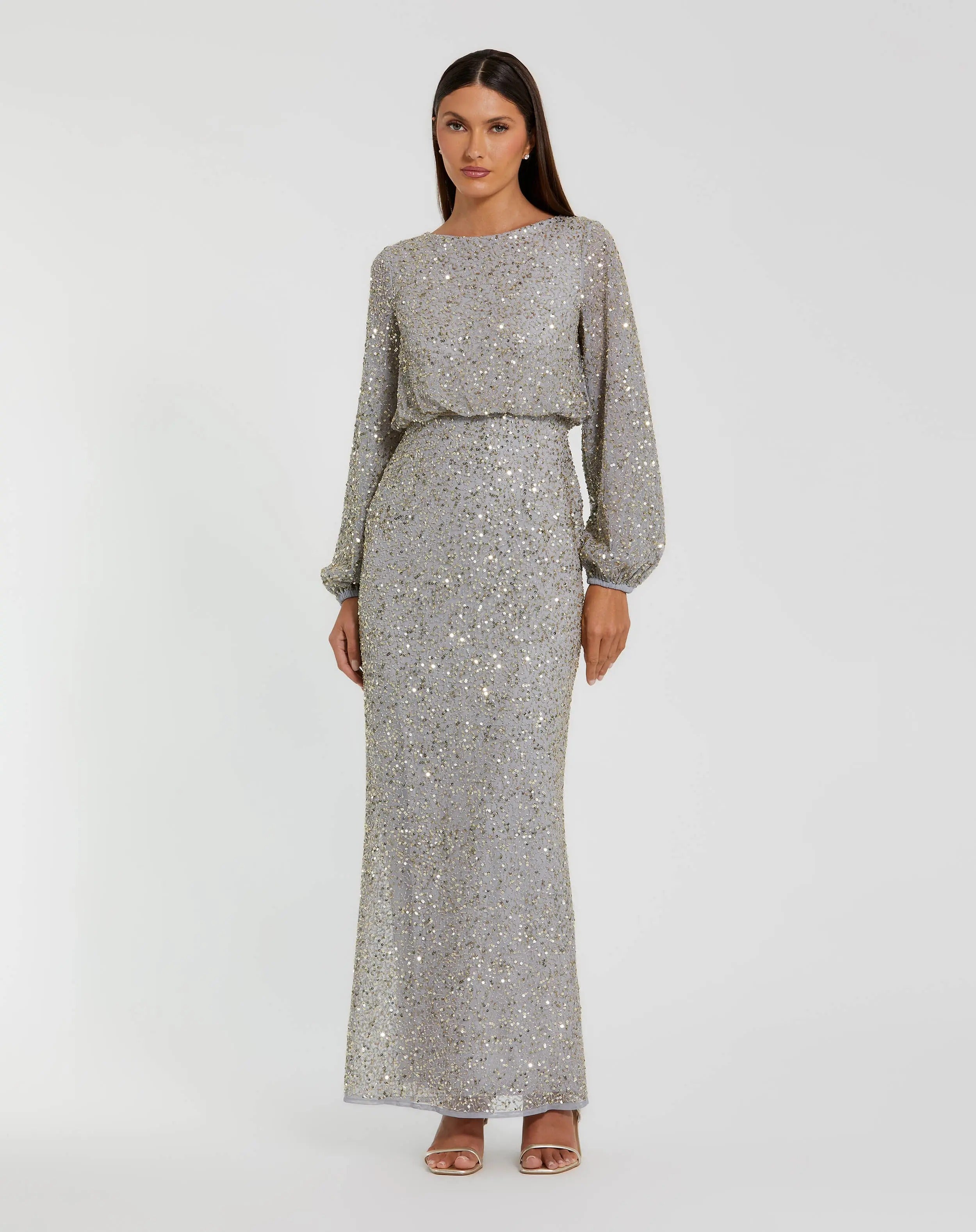 A woman stands in a full-length, long-sleeve platinum gold gown—Mac Duggal 94203—with a cinched waist and open-toe heels, set against a plain white background. This elegant sequin dress from Mac Duggal exudes both glamour and the sophistication of a modest long formal dress.